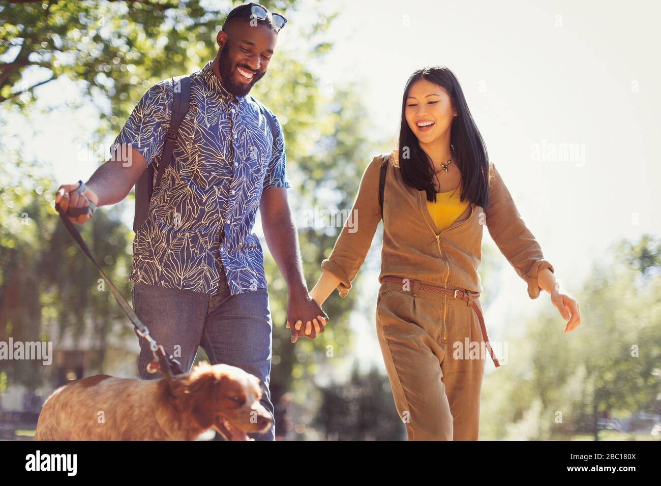 Smiling young couple walking dog in sunny park Stock Photo