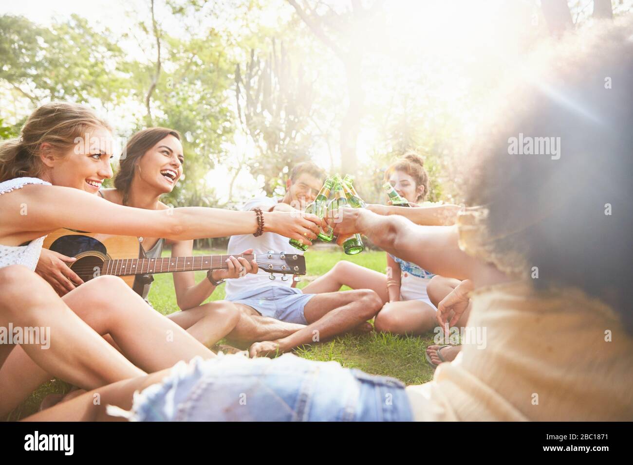 Enthusiastic young friends playing guitar and toasting beer bottles in sunny summer park Stock Photo