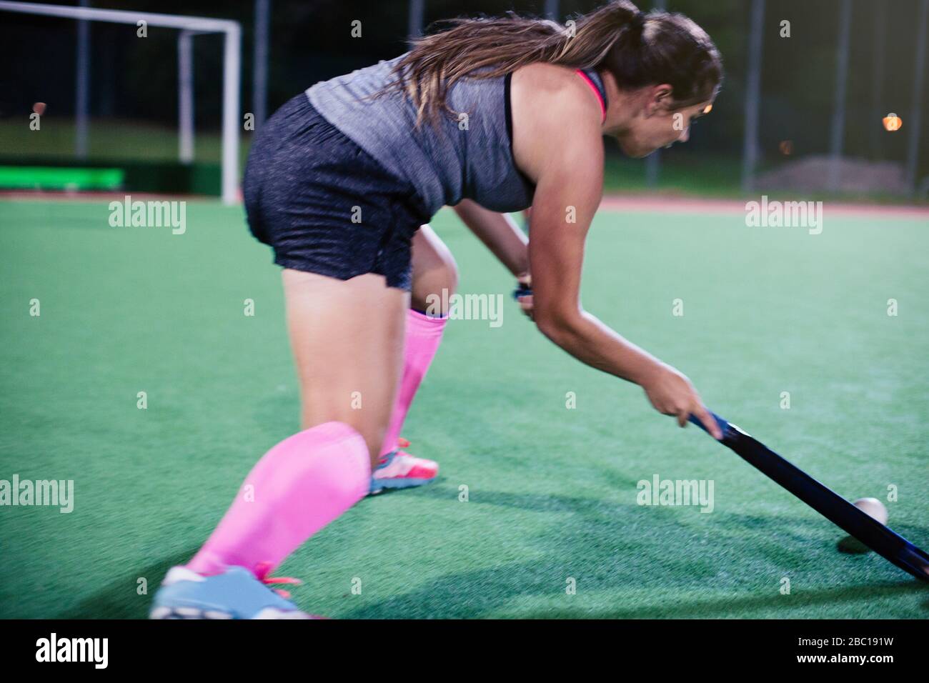 Determined young female field hockey player hitting the ball, playing on field at night Stock Photo