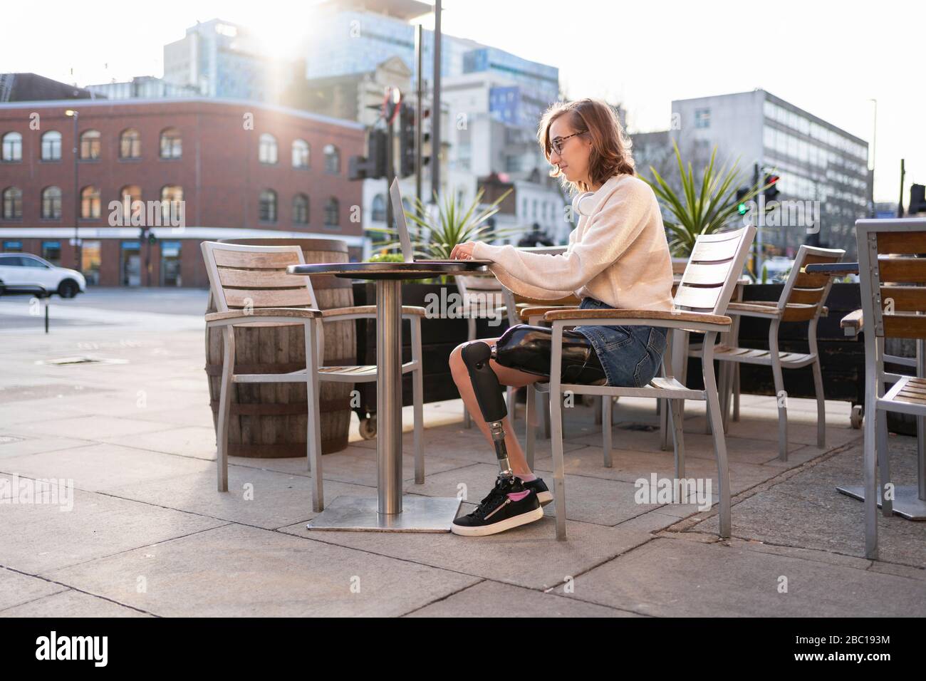 Young woman with leg prosthesis sitting in a sidewalk cafe in the city using laptop Stock Photo