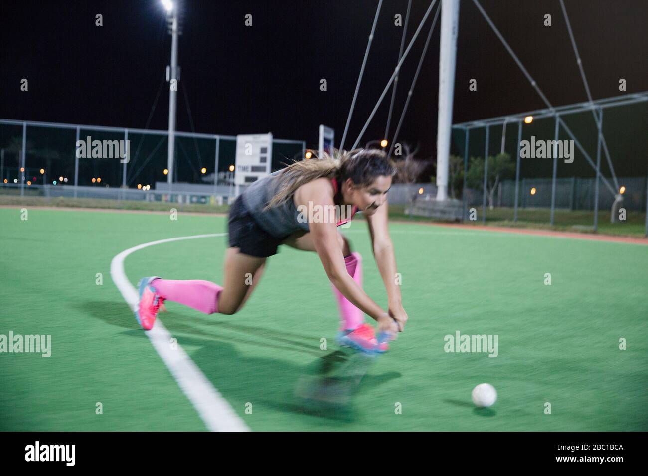 Determined young female field hockey player running for the ball, playing on field at night Stock Photo