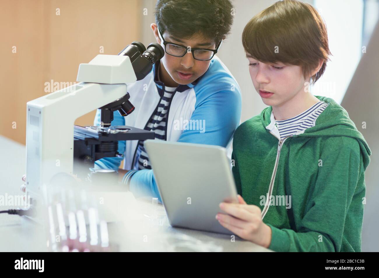 Focused boy students using digital tablet at microscope in laboratory classroom Stock Photo