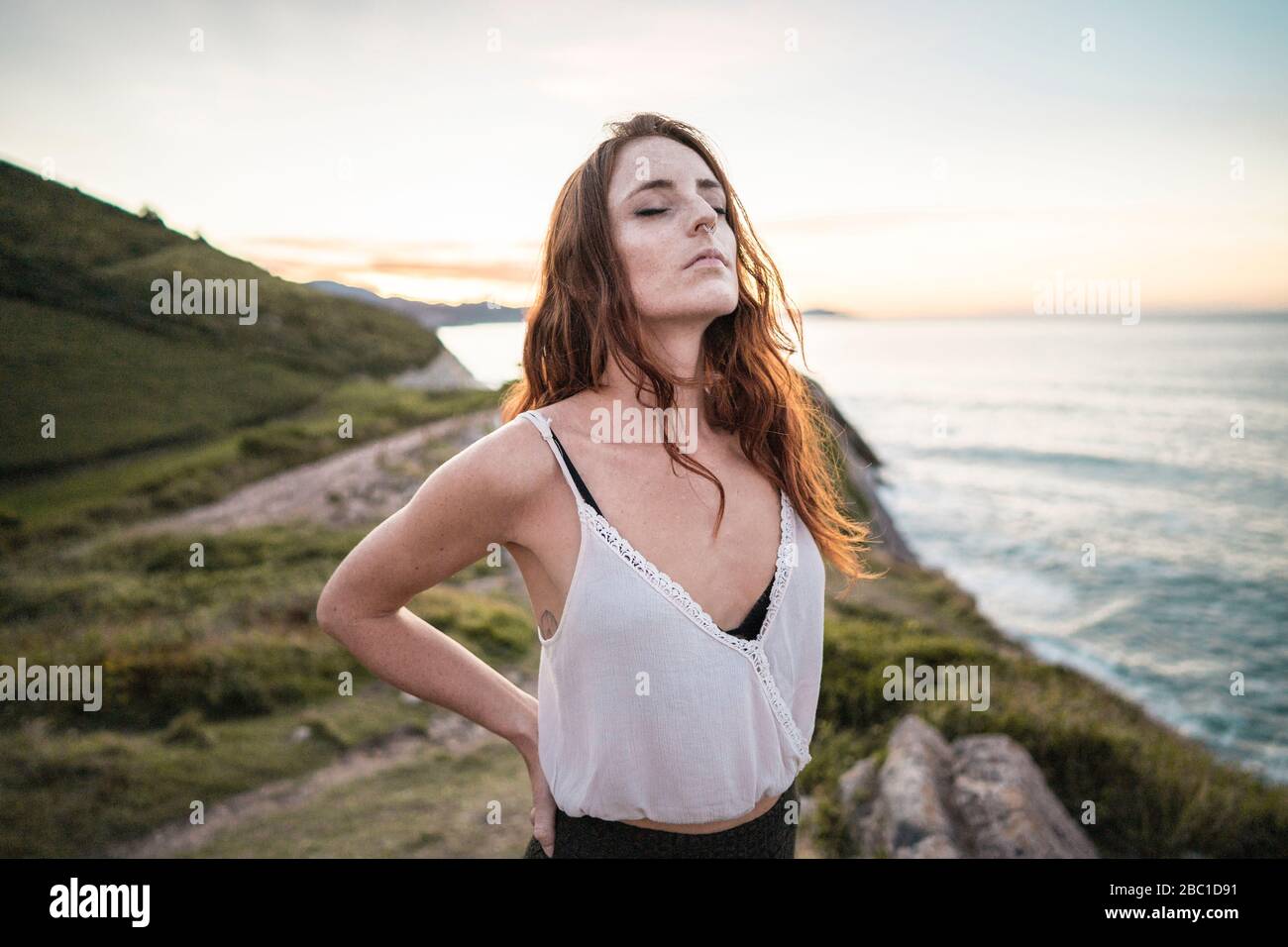 Red-haired woman with closed eyes standing on viewpoint at sunset Stock Photo