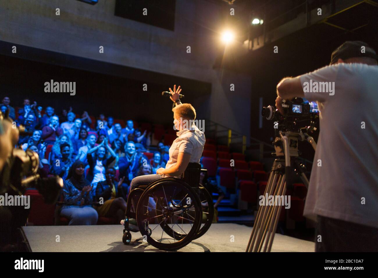 Female speaker in wheelchair on stage waving to audience Stock Photo
