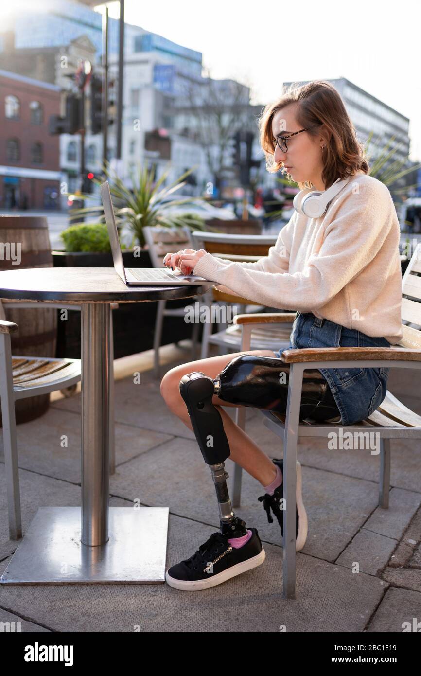 Young woman with leg prosthesis sitting in a sidewalk cafe in the city using laptop Stock Photo