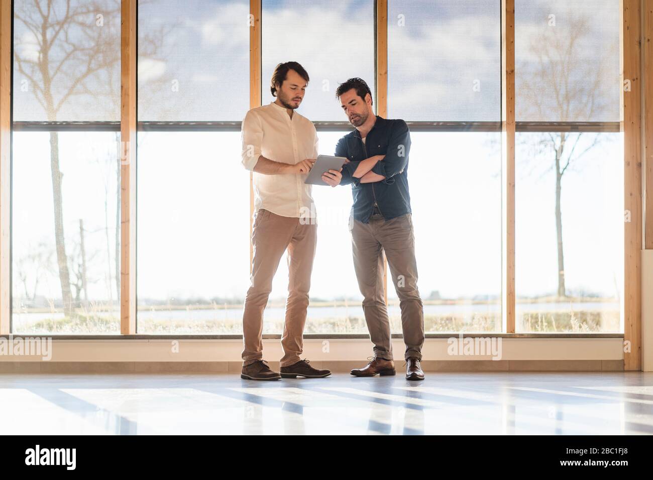 Two businessmen with tablet talking at the window in wooden open-plan ...