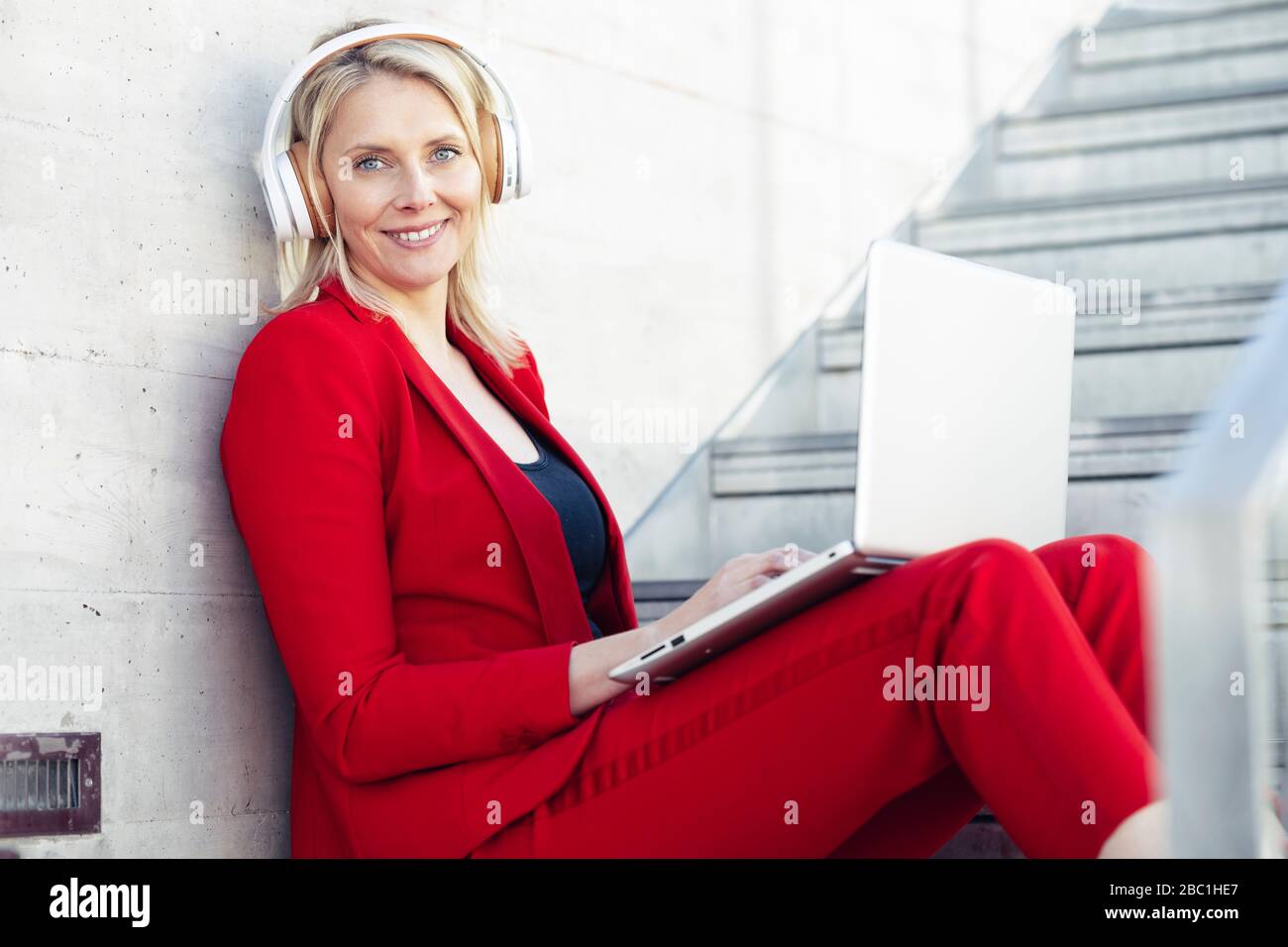 Blond businesswoman wearing red suit, sitting on stairs and using laptop Stock Photo
