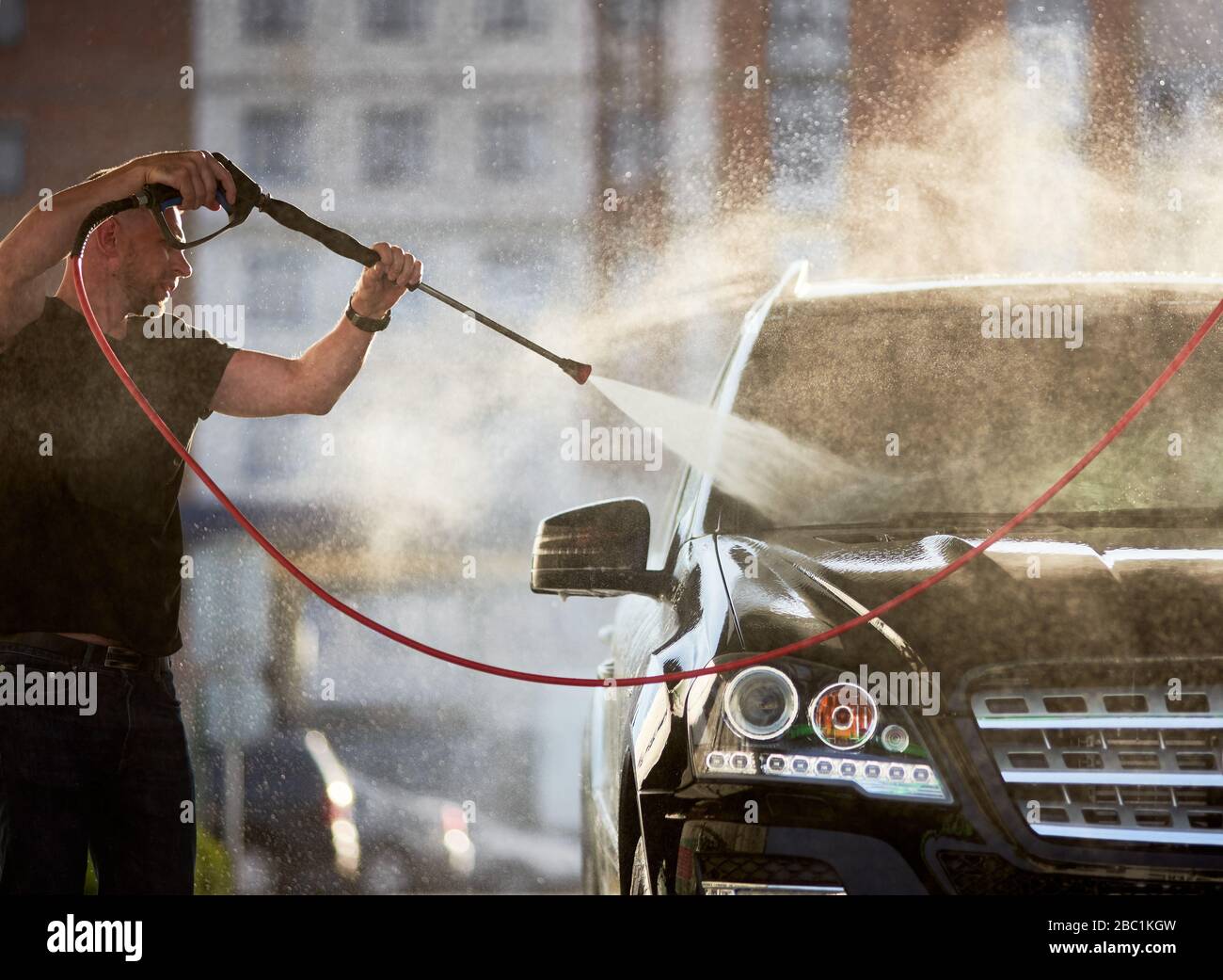Side view of man cleaning his black car outdoors, water is splashing over the top of the car shining on the sun. Stock Photo