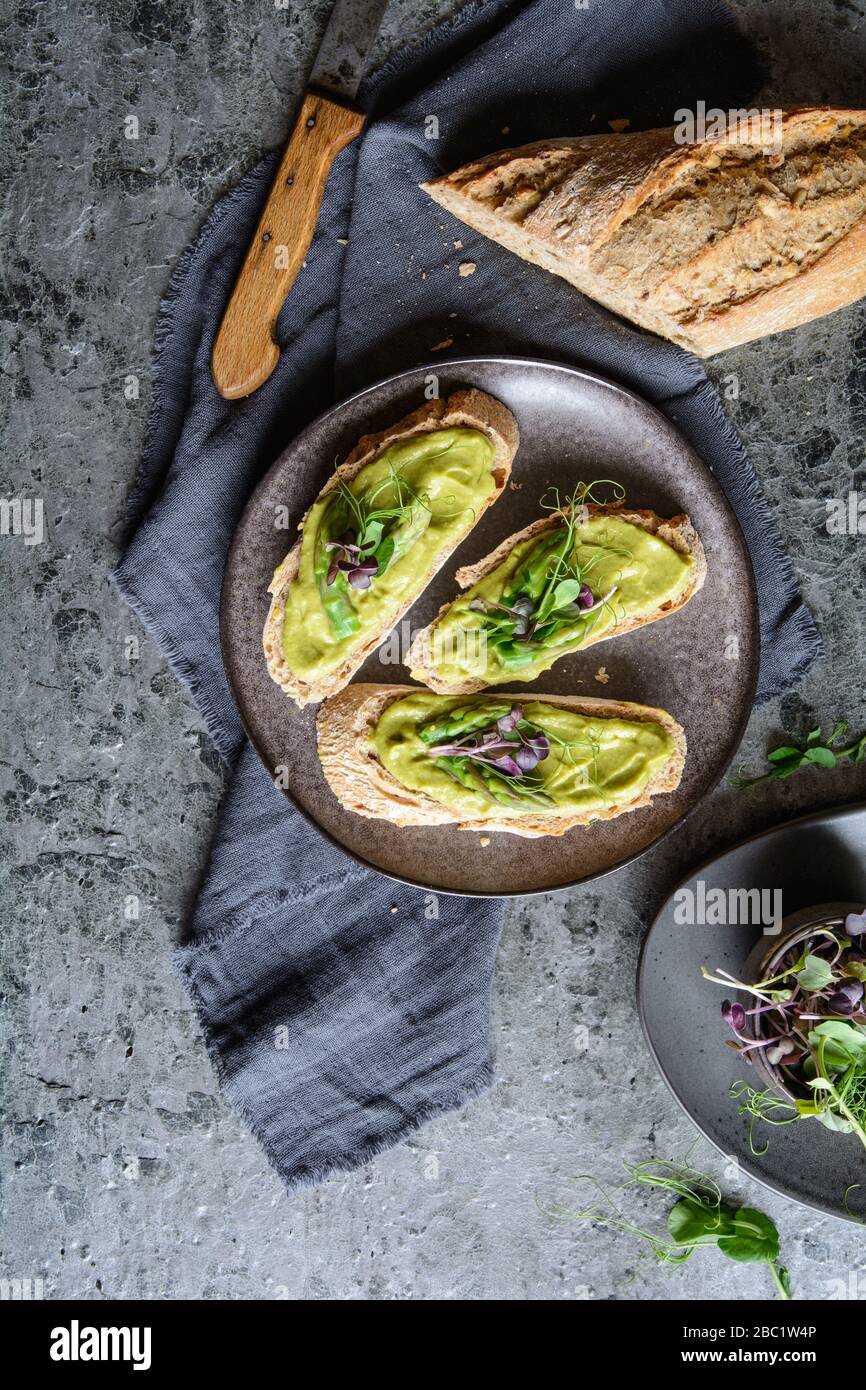 Slices of bread with vegetarian avocado, zucchini and asparagus spread, topped with pea and radish sprouts on a ceramic plate Stock Photo