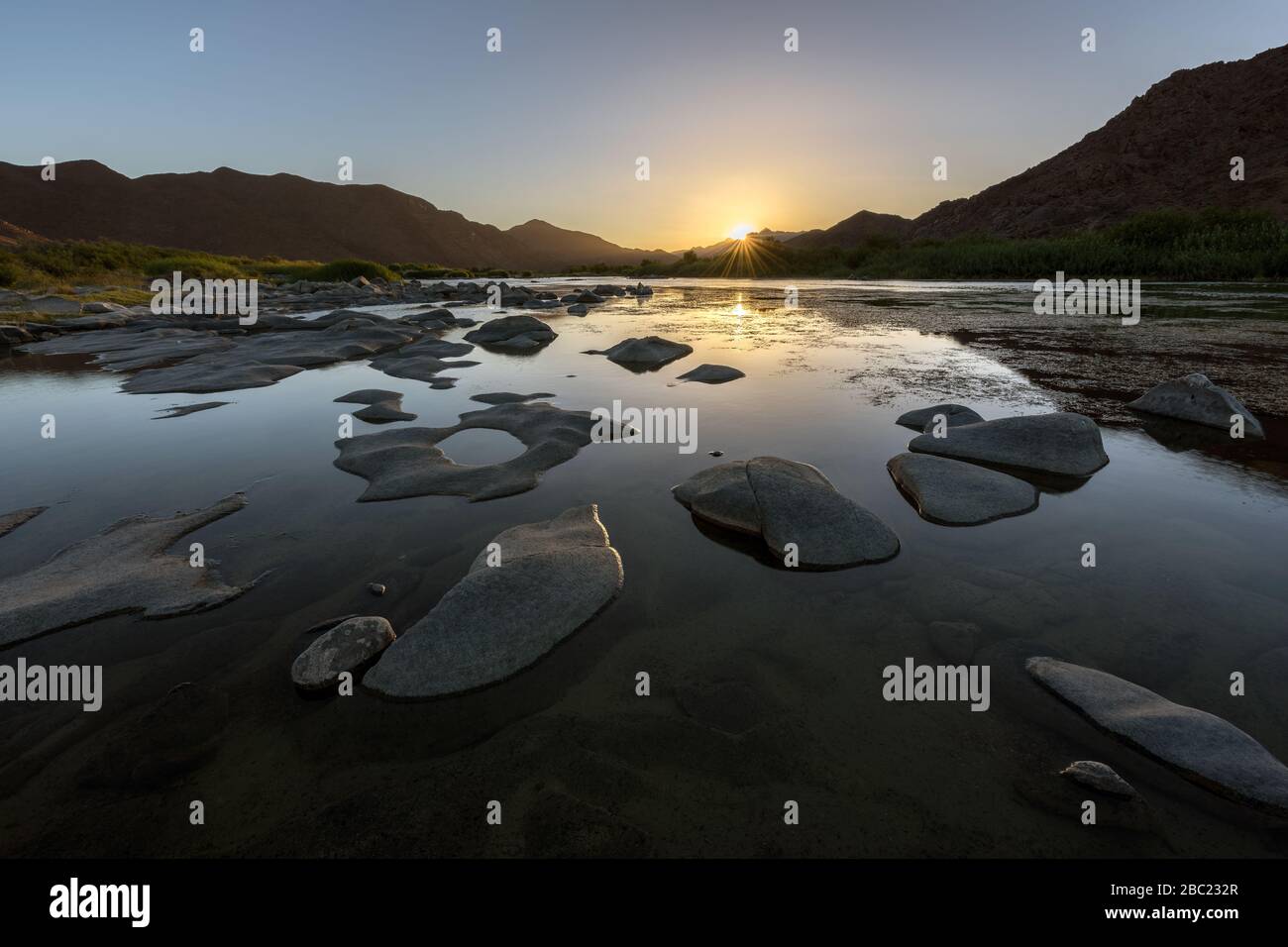 A beautiful landscape of the golden sun rising over the mountains and calm waters of the Orange River, creating a sunburst, in the Richtersveld Nation Stock Photo