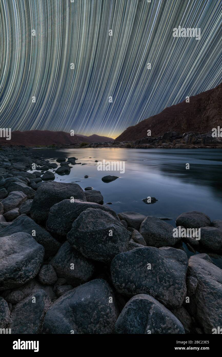 A beautiful vertical night sky landscape on the Orange River with star trails and a mountain range, a deep blue sky and rocks framing the river in the Stock Photo