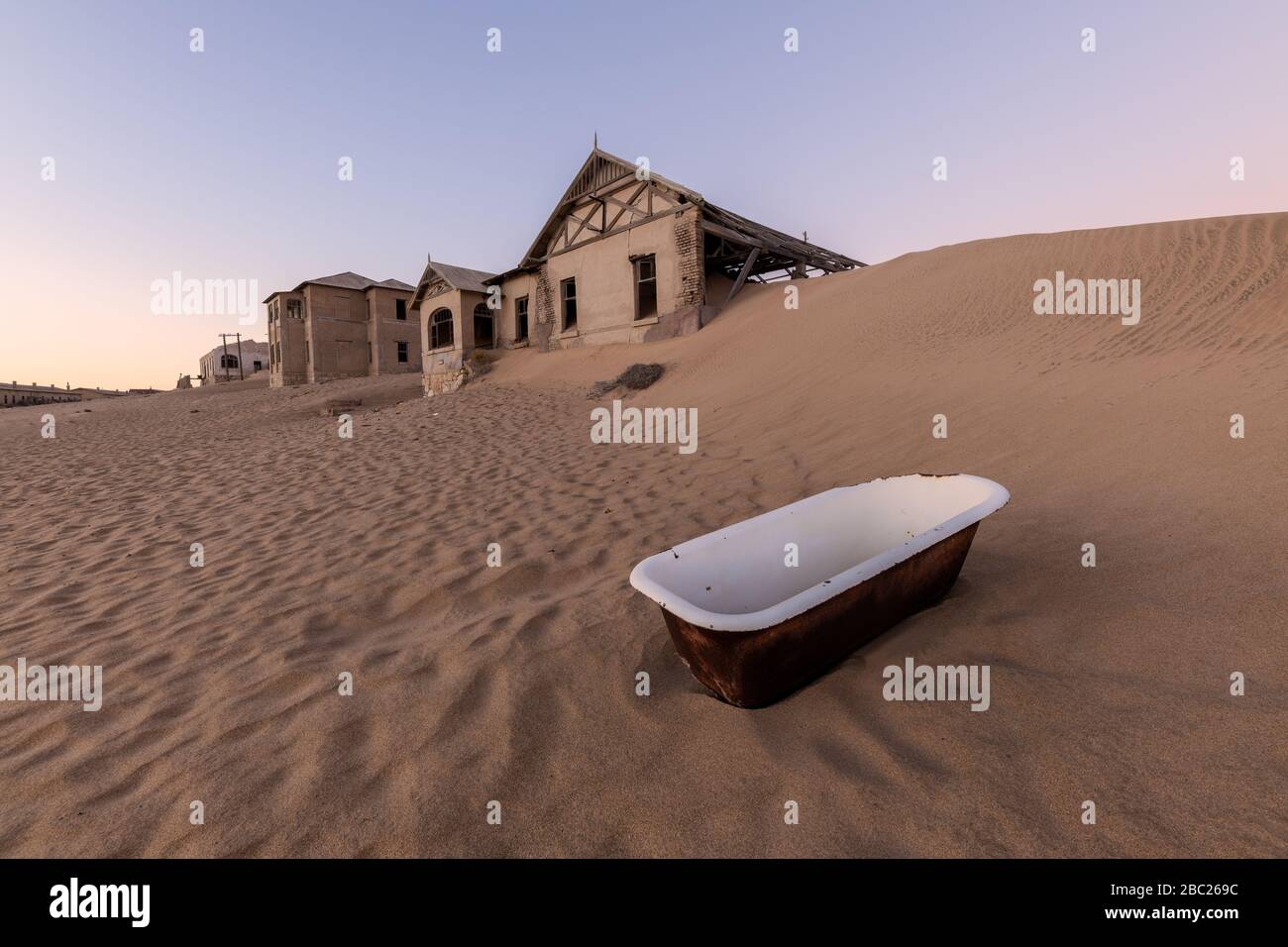 A photograph outside with an abandoned house on the horizon and a white bathtub lying in the rippled desert sand in the foreground, taken in the ghost Stock Photo