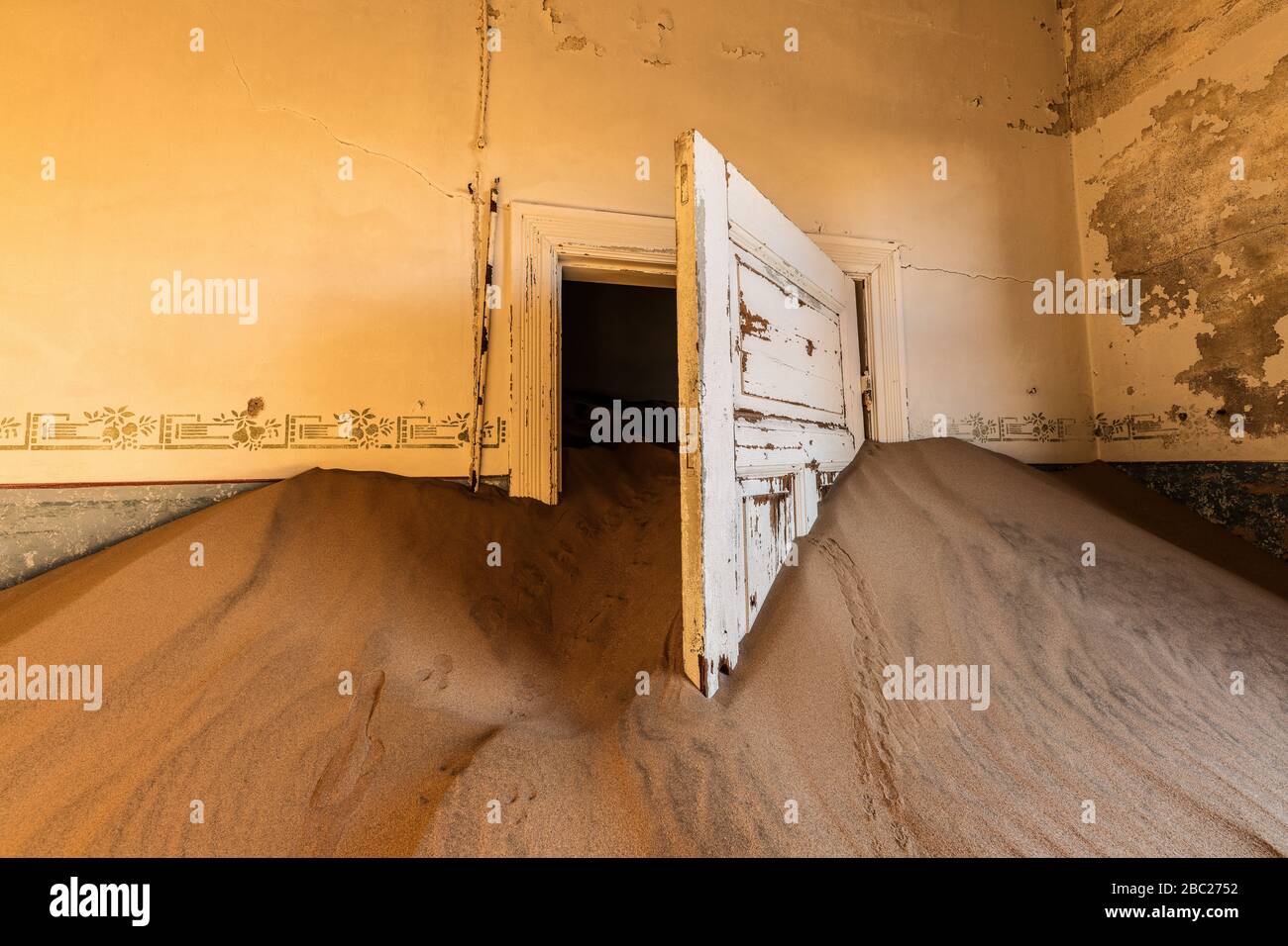A photograph inside an abandoned house with an open white door submerged in the rippled desert sand, taken in the ghost town of Kolmanskop, Namibia. Stock Photo