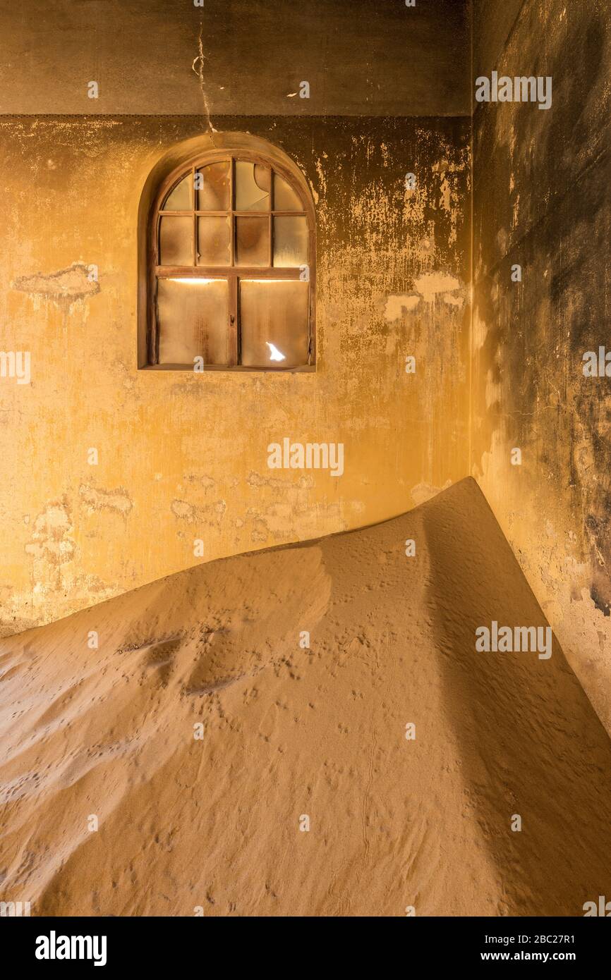 A vertical photograph inside an abandoned house with desert sand piled in the corner and a broken window in the wall, taken in the ghost town of Kolma Stock Photo