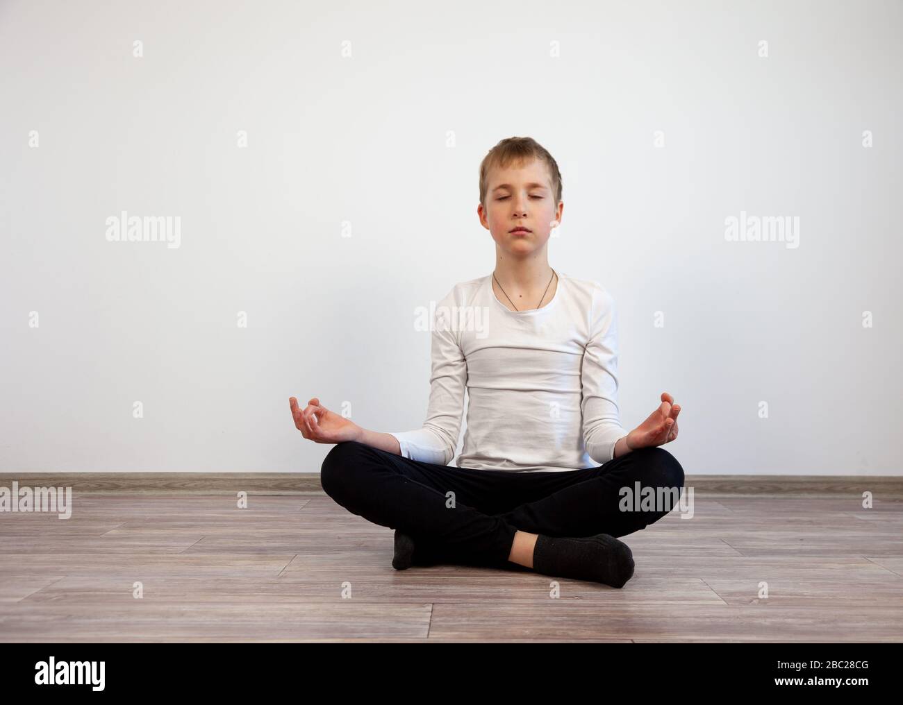 The boy is meditating at home sitting on the floor. Teenager do meditation. Self-isolation, calm, yoga. Lotus position. Exercises in the room. Stock Photo