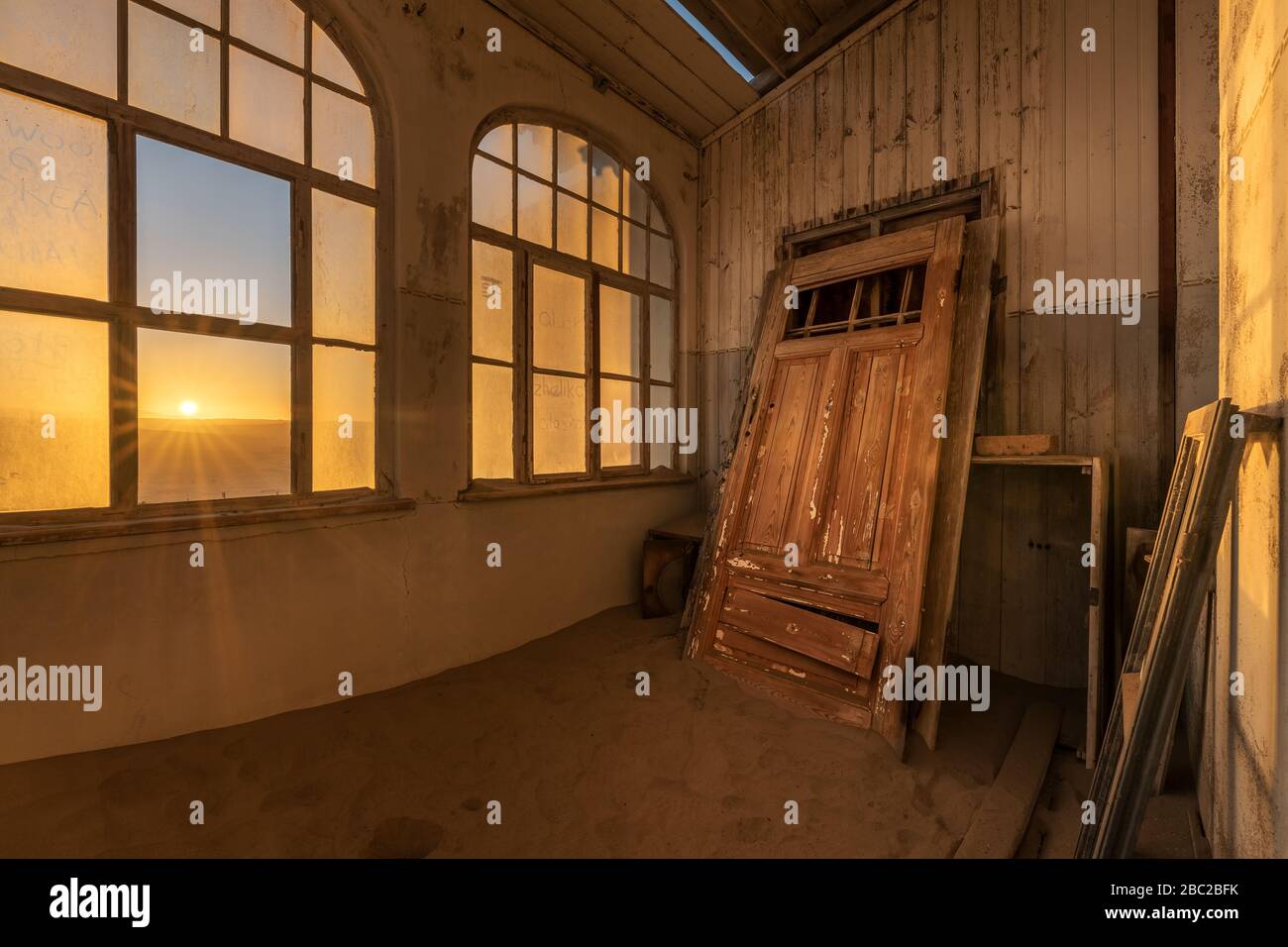 A dramatic photograph inside an abandoned house at sunrise, with a golden sunburst through broken windows and an ancient door buried in desert sand, t Stock Photo