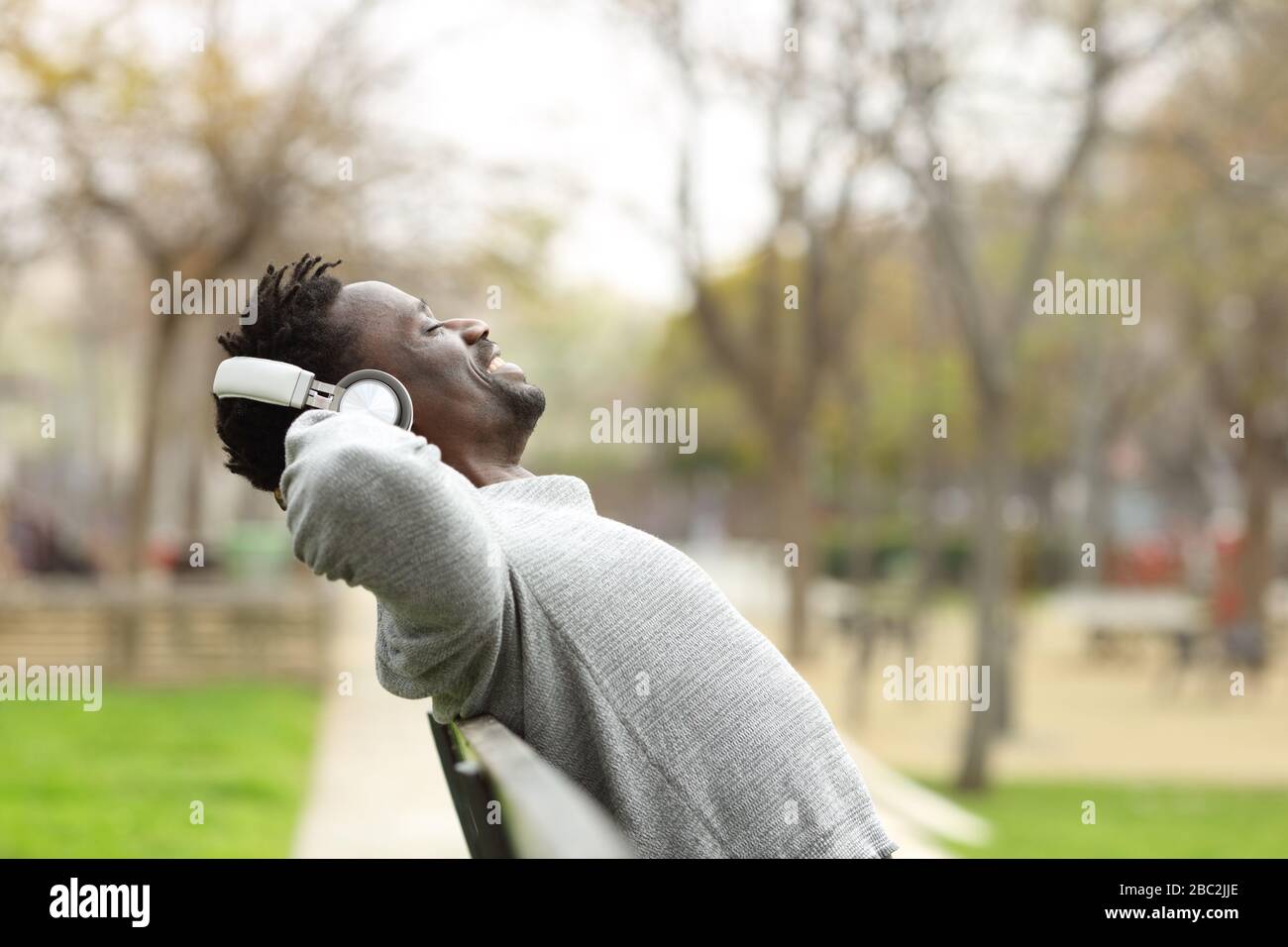 Side view portrait of a black man wearing wireless headphones relaxing and listening to music sitting on a on a bench in a park Stock Photo