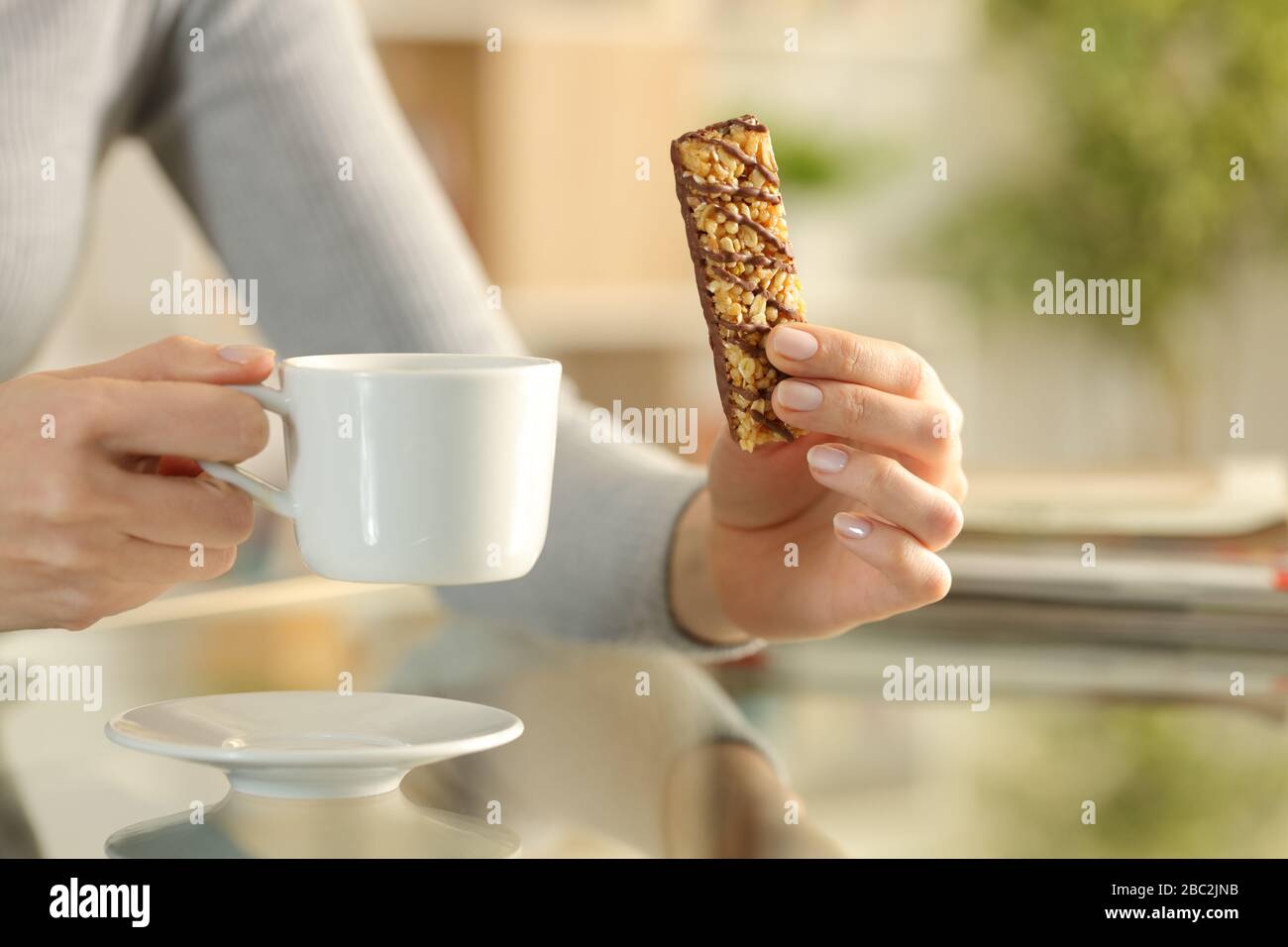 Close up of woman hands holding a snack bar and coffee cup sitting on a desk at home at breakfast Stock Photo