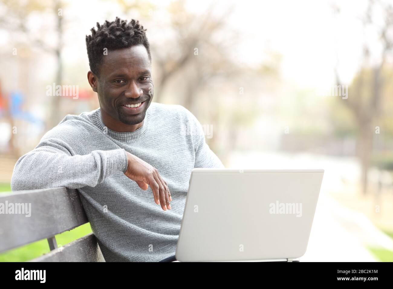 Happy black man with laptop looking at camera sitting on a park bench Stock Photo