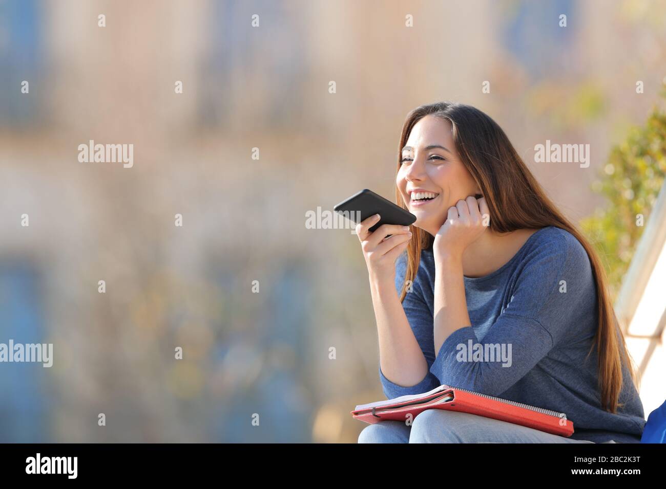 Happy student using voice recognition on mobile phone sitting outdoors in a university campus Stock Photo