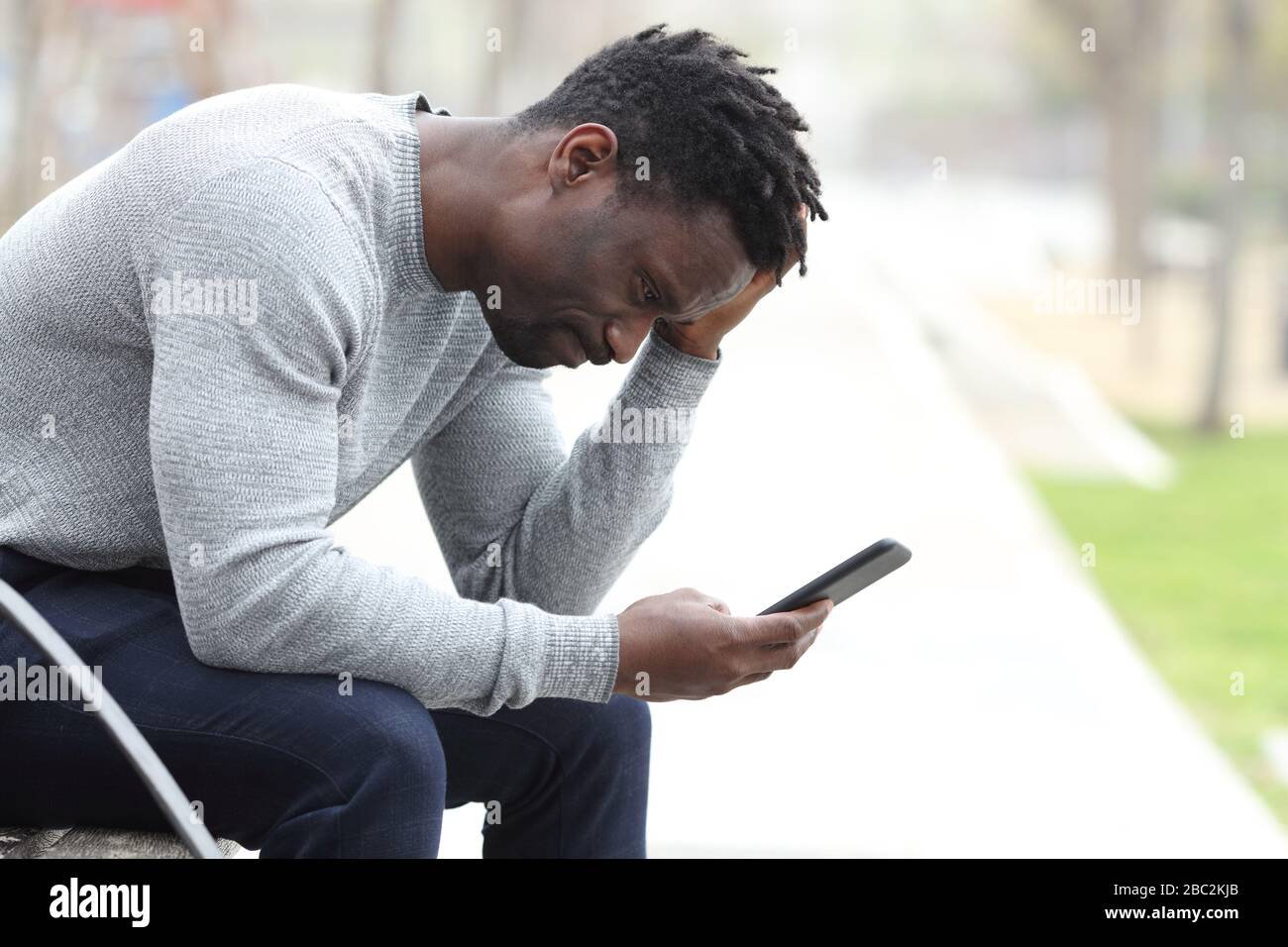 Side view portrait of a sad black man complaining checking mobile phone sitting on a bench in a park Stock Photo