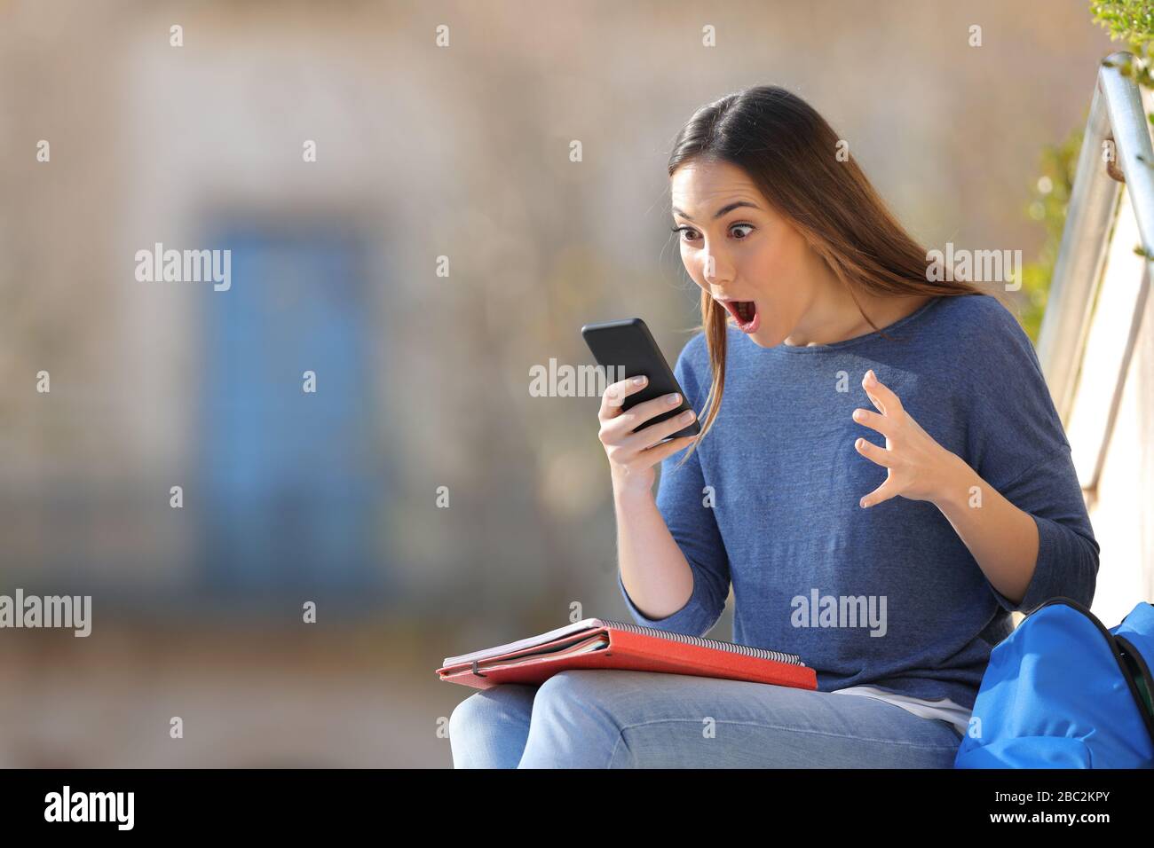 Surprised student checking mobile phone sitting outdoors in a university campus Stock Photo