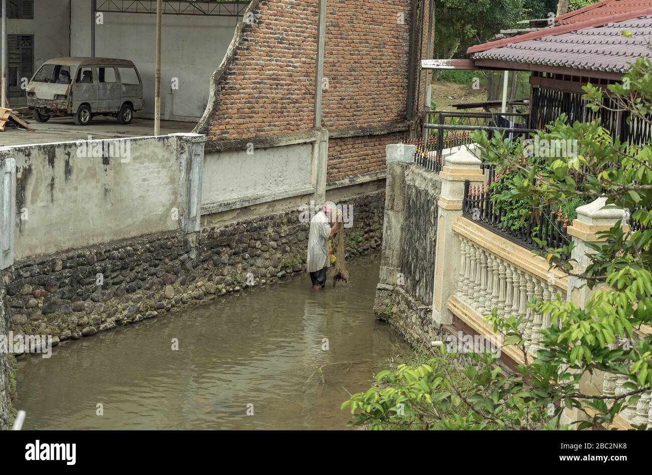 21 June 2018 Panyambungan,Sumatra, Indonesia: Local poor man fishing with net in murky dirty river in middle of town also old dirty shabby car visible Stock Photo
