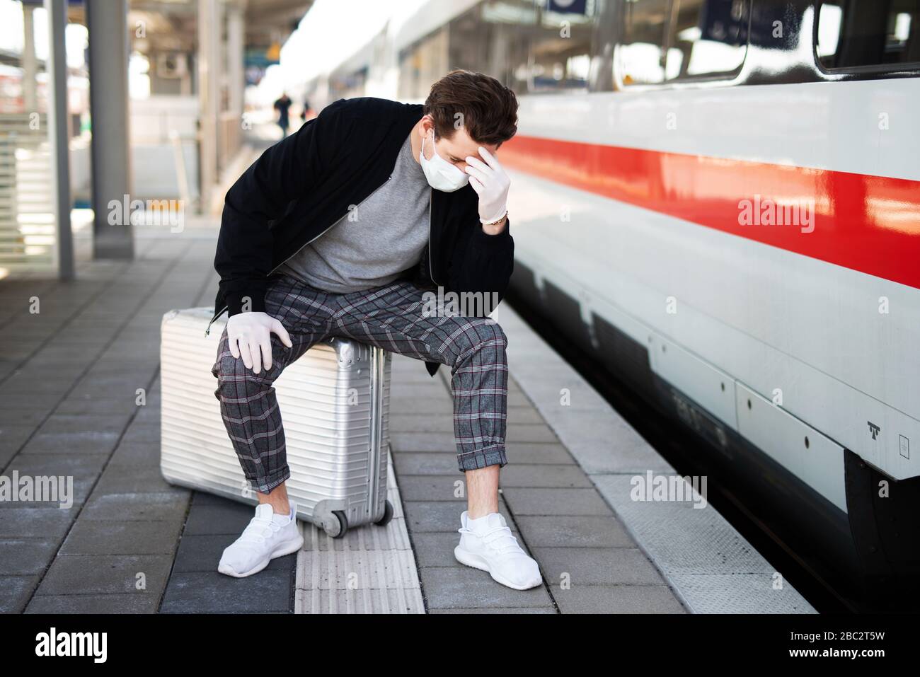 Man with a suitcase on the platform, he has a face mask and gloves against infection with the corona virus Stock Photo