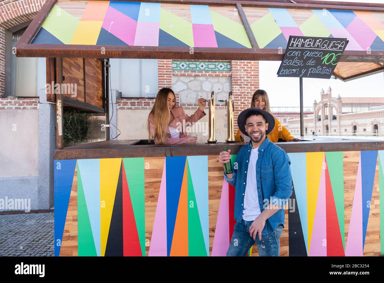 Two young blond-haired Caucasian women working at an outdoor bar toast and celebrate with a young Hispanic customer wearing a hat Stock Photo