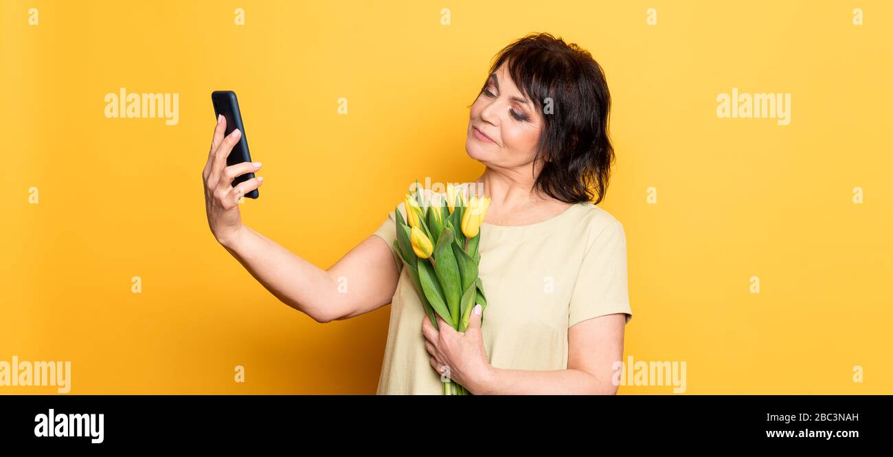Beautiful old woman with professional smokey make-up and hairstyle holding tulip flowers on the pink background. Taking video call by the smartphone Stock Photo