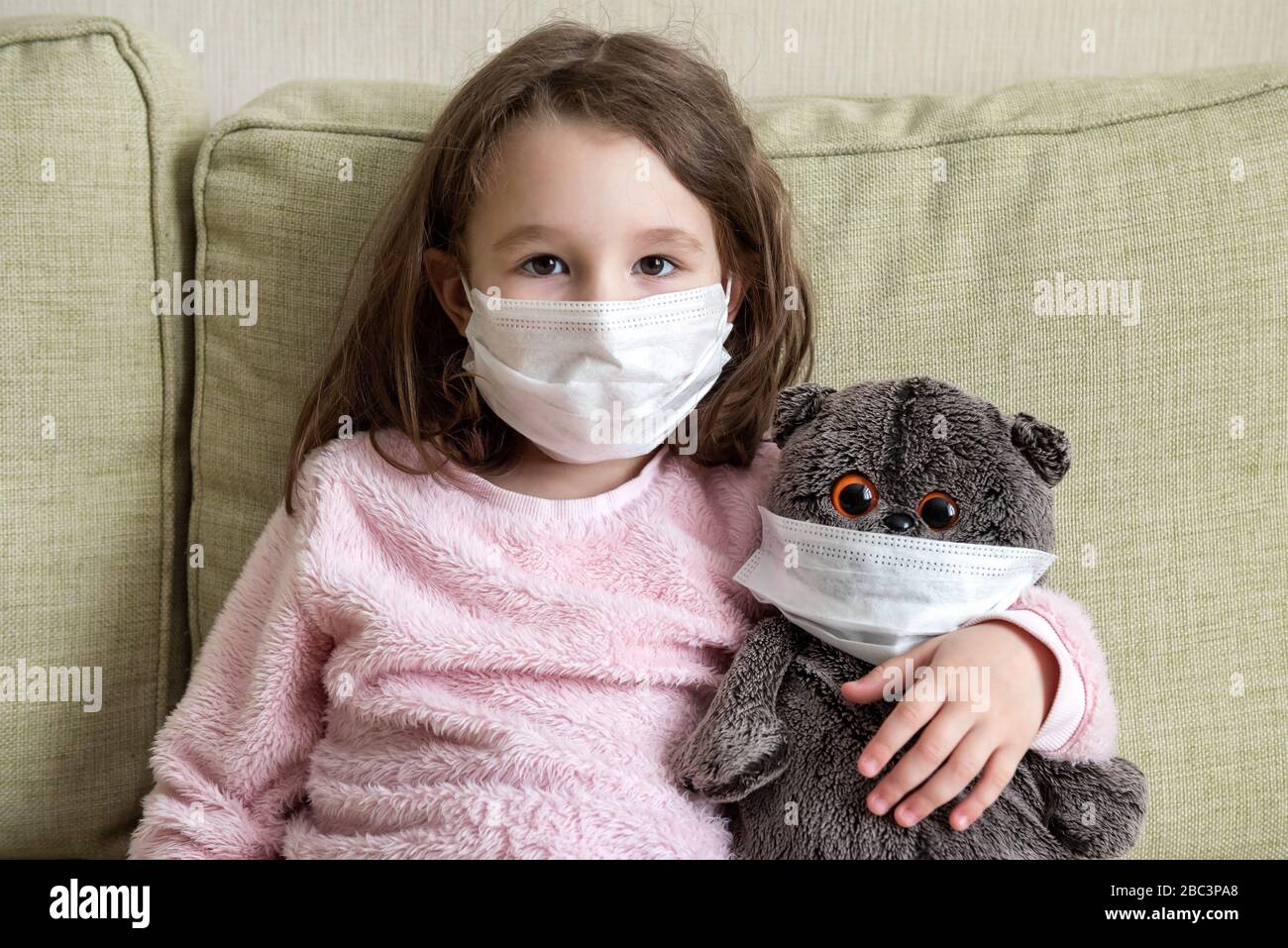 Stay at home in quarantine, funny kid with toy on couch during COVID-19 coronavirus pandemic. Little girl in medical mask for protection to corona vir Stock Photo