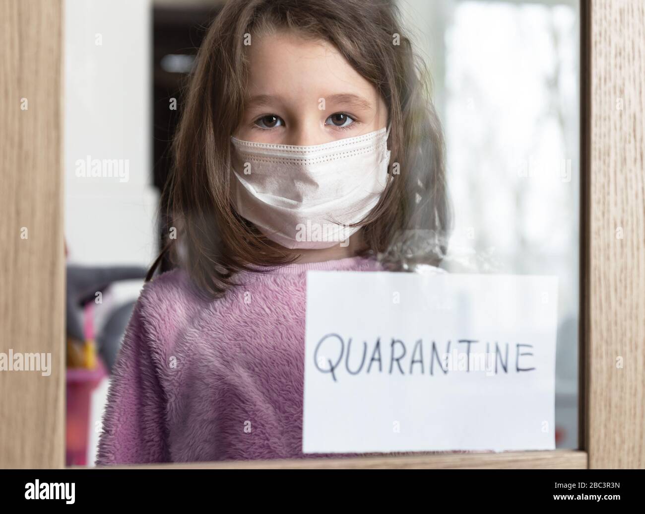 COVID-19 coronavirus concept, little girl in medical mask looking through window with note Quarantine at home. Sad kid during quarantine due to COVID Stock Photo