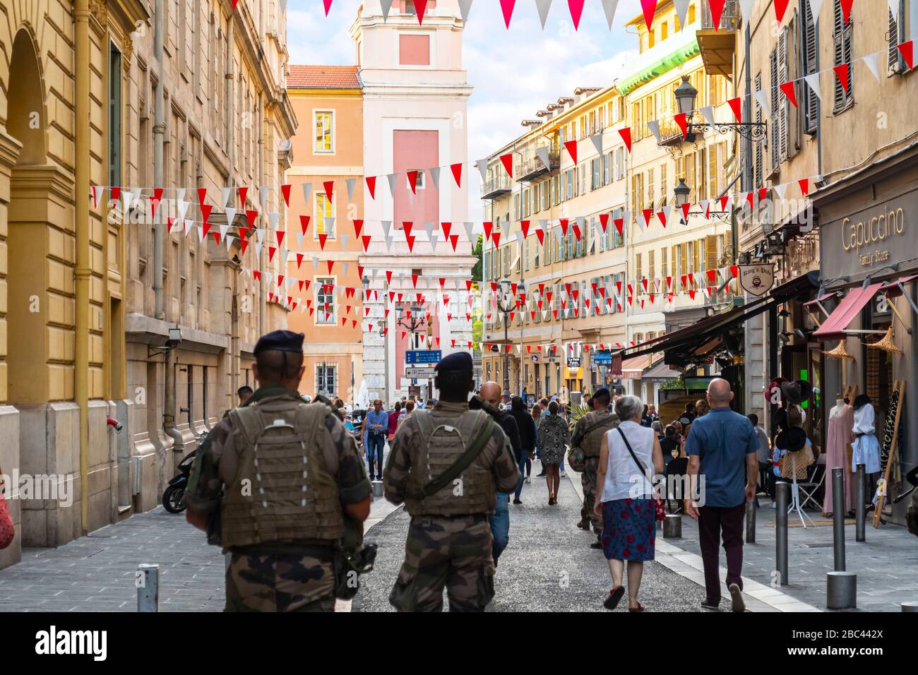 Military security armed police walk through Old Vieux Nice, France, towards Rusca Palace as flags drape across the alley late in the day. Stock Photo
