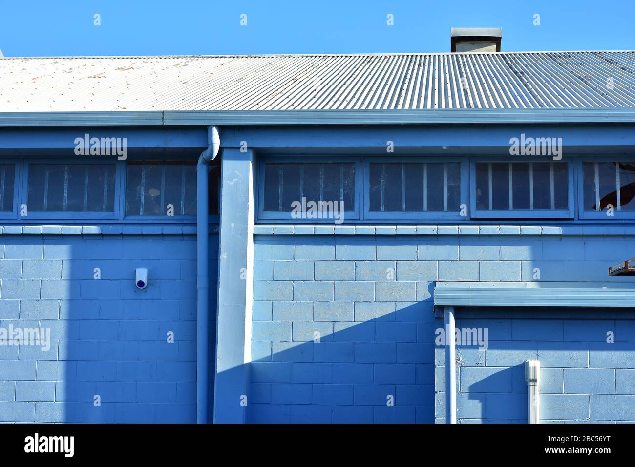 Back side of industrial building painted pale blue with windows with metal bars and corrugated metal roof. Stock Photo