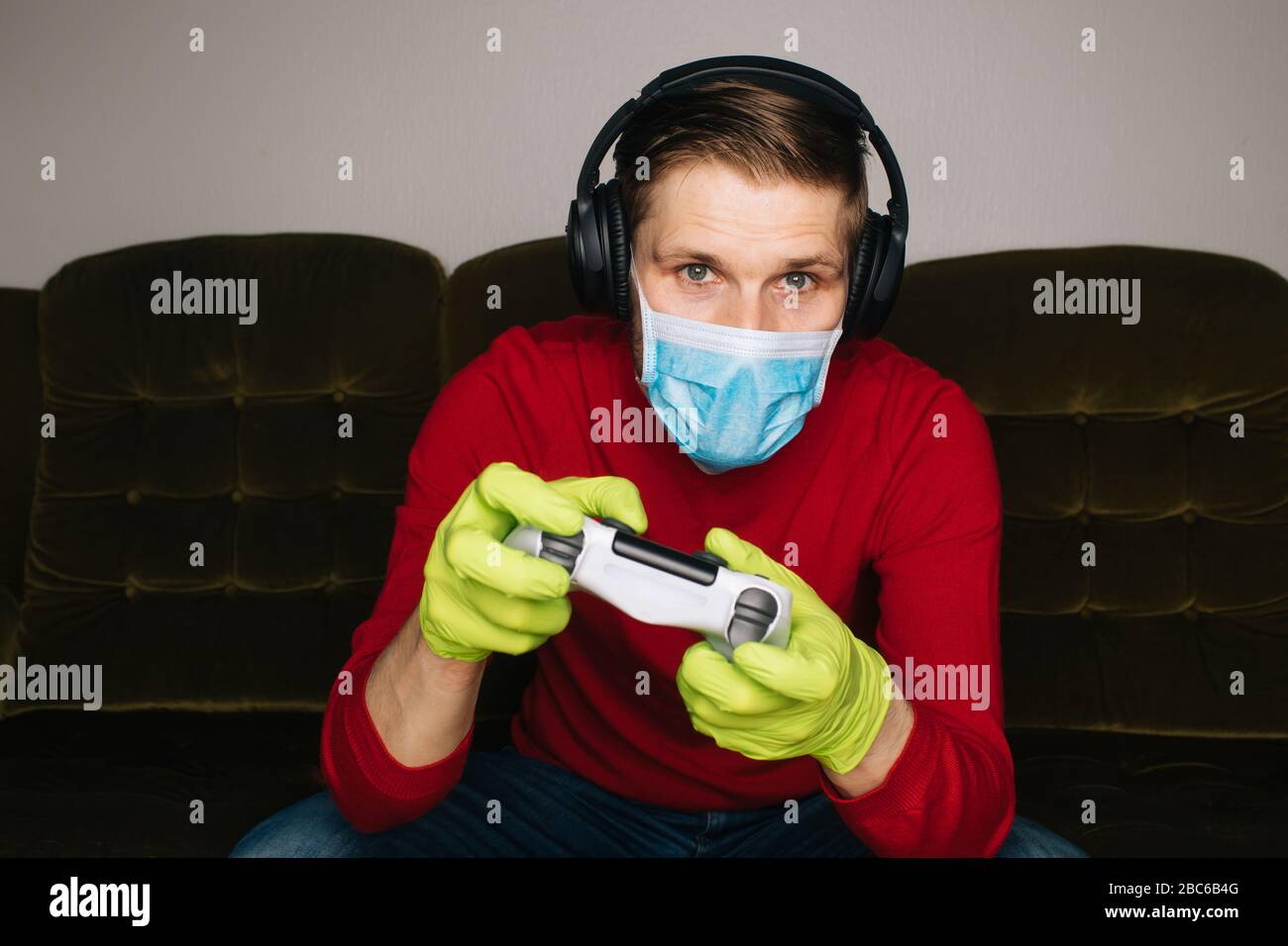 Man playing video game wearing a surgical mask, rubber gloves and noise cancelling headphones. Self-isolation (self-quarantine) for coronavirus (COVID Stock Photo
