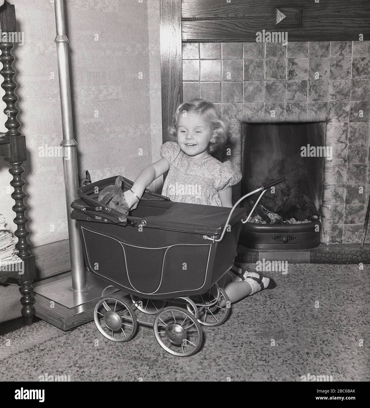 1950s, in a living room, in front of an open coal fire, a young girl playing with her soft toy sat in a small four-wheeled doll's pram of the era, England, UK. Stock Photo