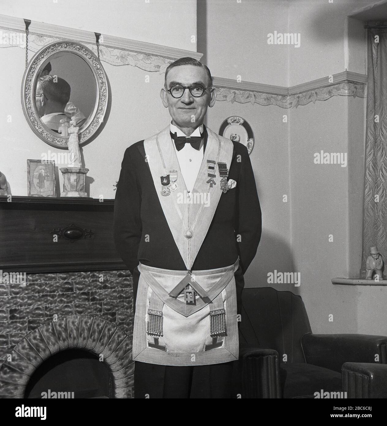 1950s, historical, Standing proudly by the fireplace in his front room, a freemason wearing his dress apron or regalia. Also known as a Craft Master Masons apron, it derives from the working apron of the ancient stone masons and is considered an emblem of innocence and purity of life. A master mason's apron is normally made from lambskin and in primitive times it was an ecclesiastical (religious) decoration. Stock Photo