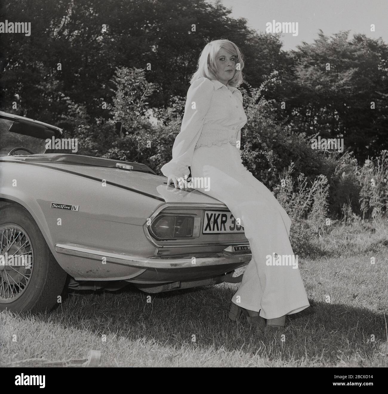 1970s, historical, Outside in a sun-lit field, an attractive blonde woman in a white top and bell-bottomed trousers, standing beside her open-top sports car, a Triumph Spitfire IV, with wire wheels, England, UK. First introduced in 1962, based on a design by Giovanni Michelotti, the Spitfire was a small British two-seat sports car and manufactured up to 1980. The mark IV model shown here was made at Standard-Triumph works (owned by Leyland Motors) in Canley, Coventry between 1970 and 1974 and among other new features from previous models, was a newly designed rear end. Stock Photo