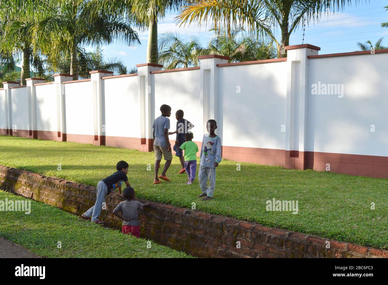 LILONGWE, MALAWI, AFRICA - APRIL 2, 2018: Local children are plaing on the ground with bright green grass near white fence and palms, one boy is smili Stock Photo