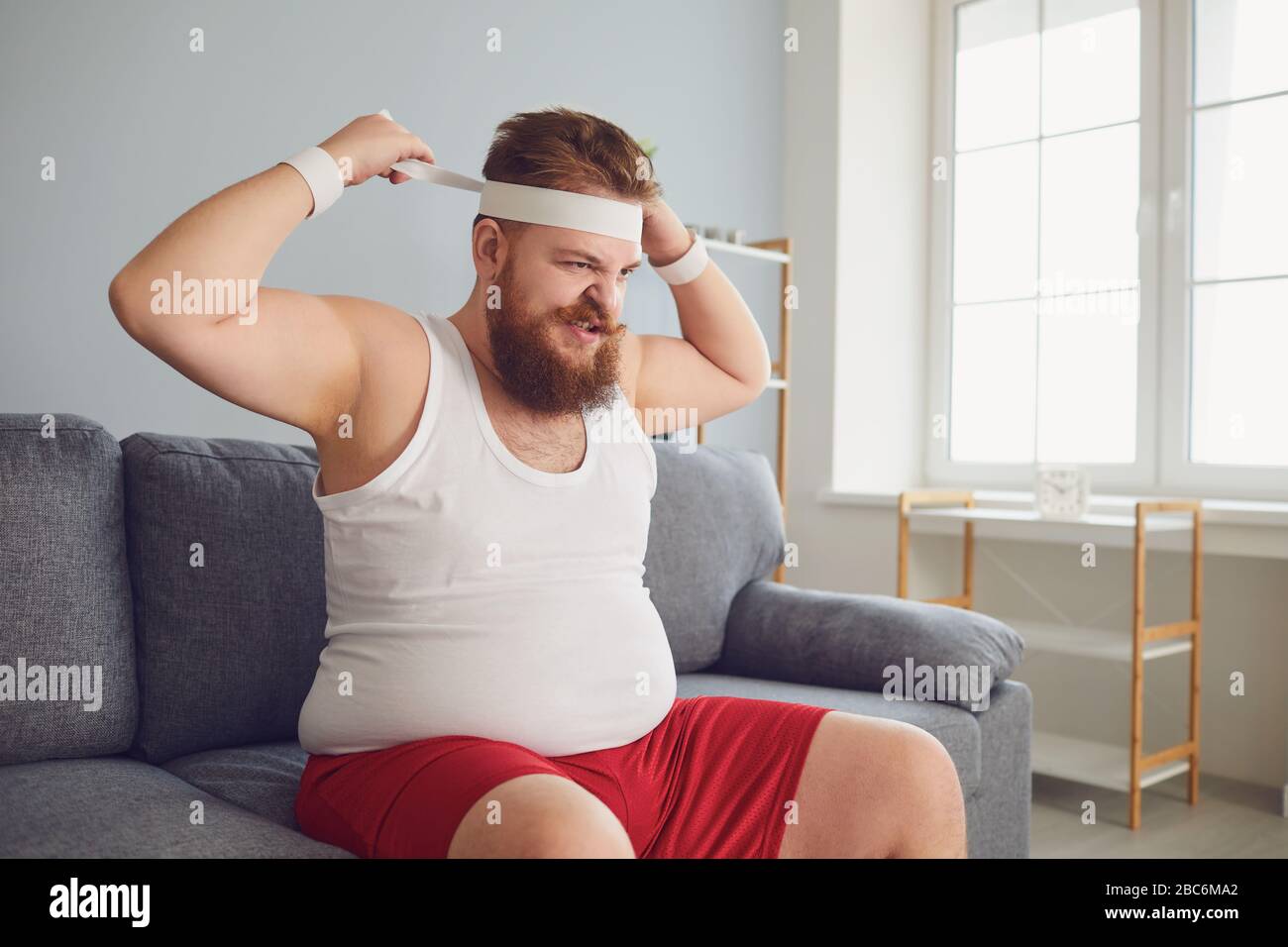 Funny fat angry man in sportswear is sitting on the sofa in the room Stock Photo