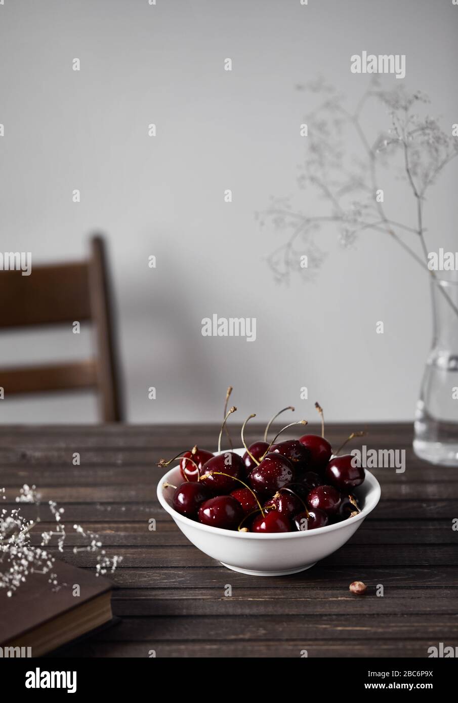 Black cherry in the white bowl on the wooden table near white flowers and book at white background in summer time Stock Photo