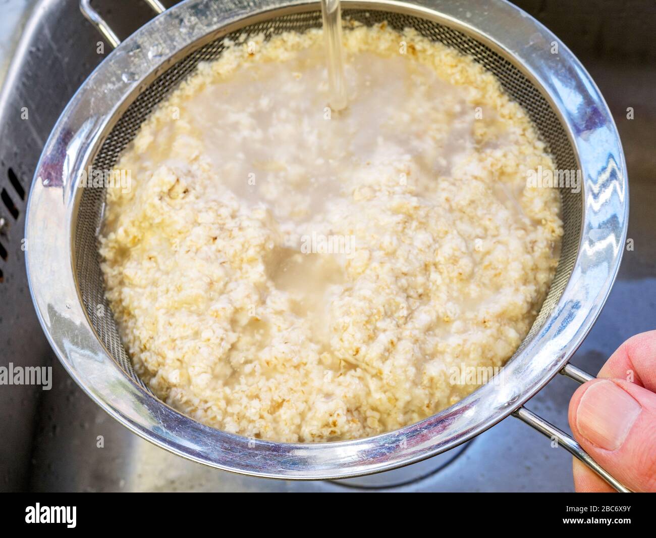 Draining and rinsing soaked rolled oats in a sieve over a kitchen sink for making dairy alternative oat cream Stock Photo