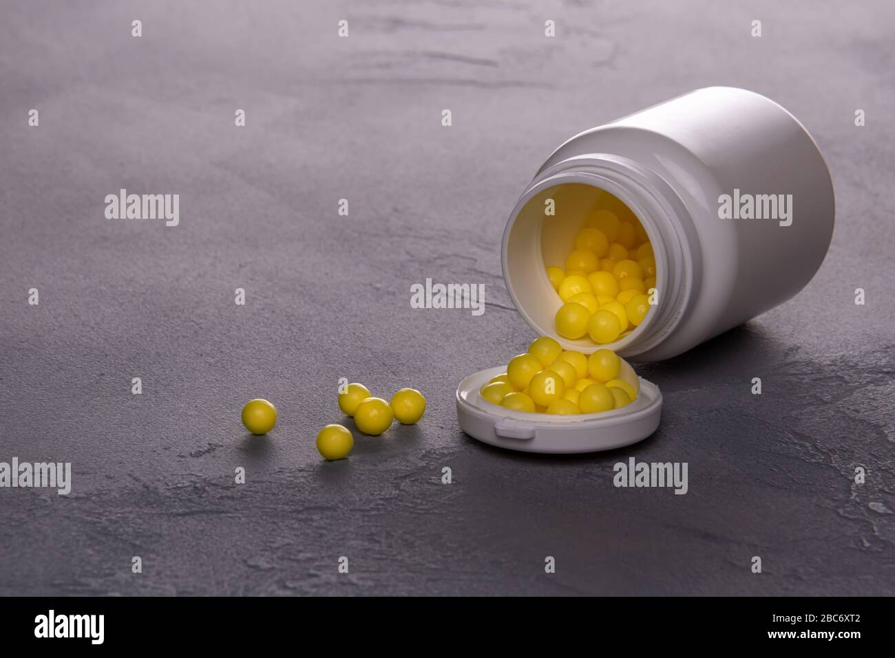 Horizontal textured gray background with opened white plastic bottle lying on side and full of small round yellow pills of Vitamin C. Few pills are sc Stock Photo