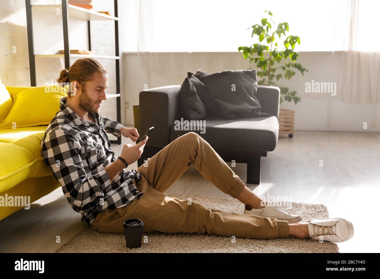 Photo of young handsome man wearing plaid shirt holding cellphone and sitting on floor in apartment Stock Photo