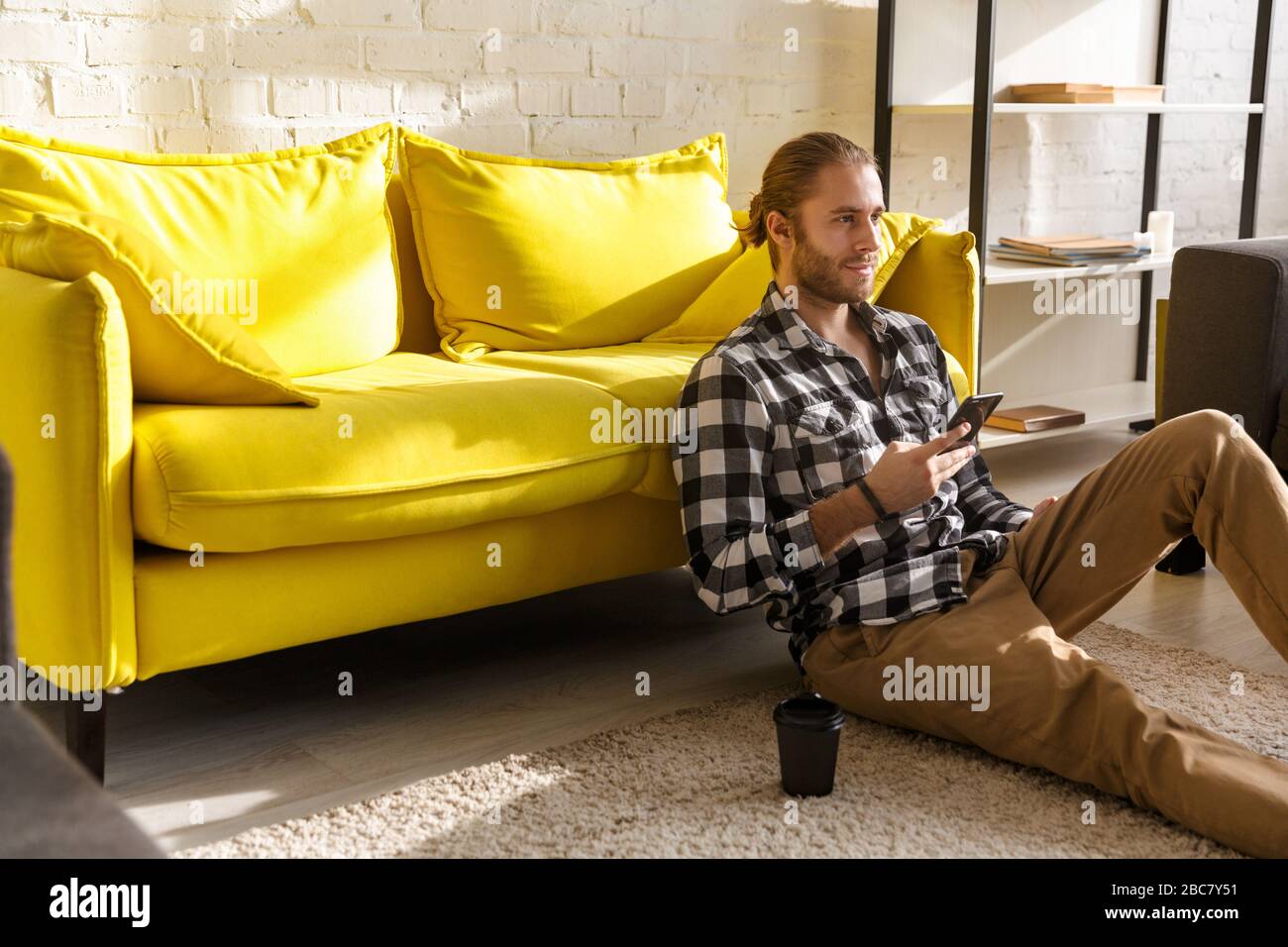 Photo of young handsome man wearing plaid shirt holding cellphone and sitting on floor in apartment Stock Photo