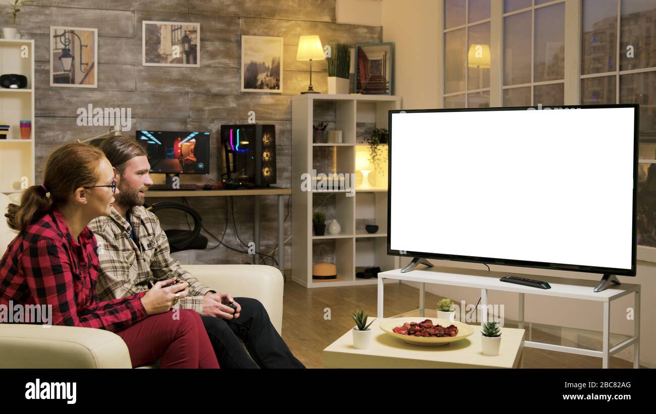 Relaxed couple sitting on sofa playing video games using wireless joysticks on tv with green screen. Stock Photo