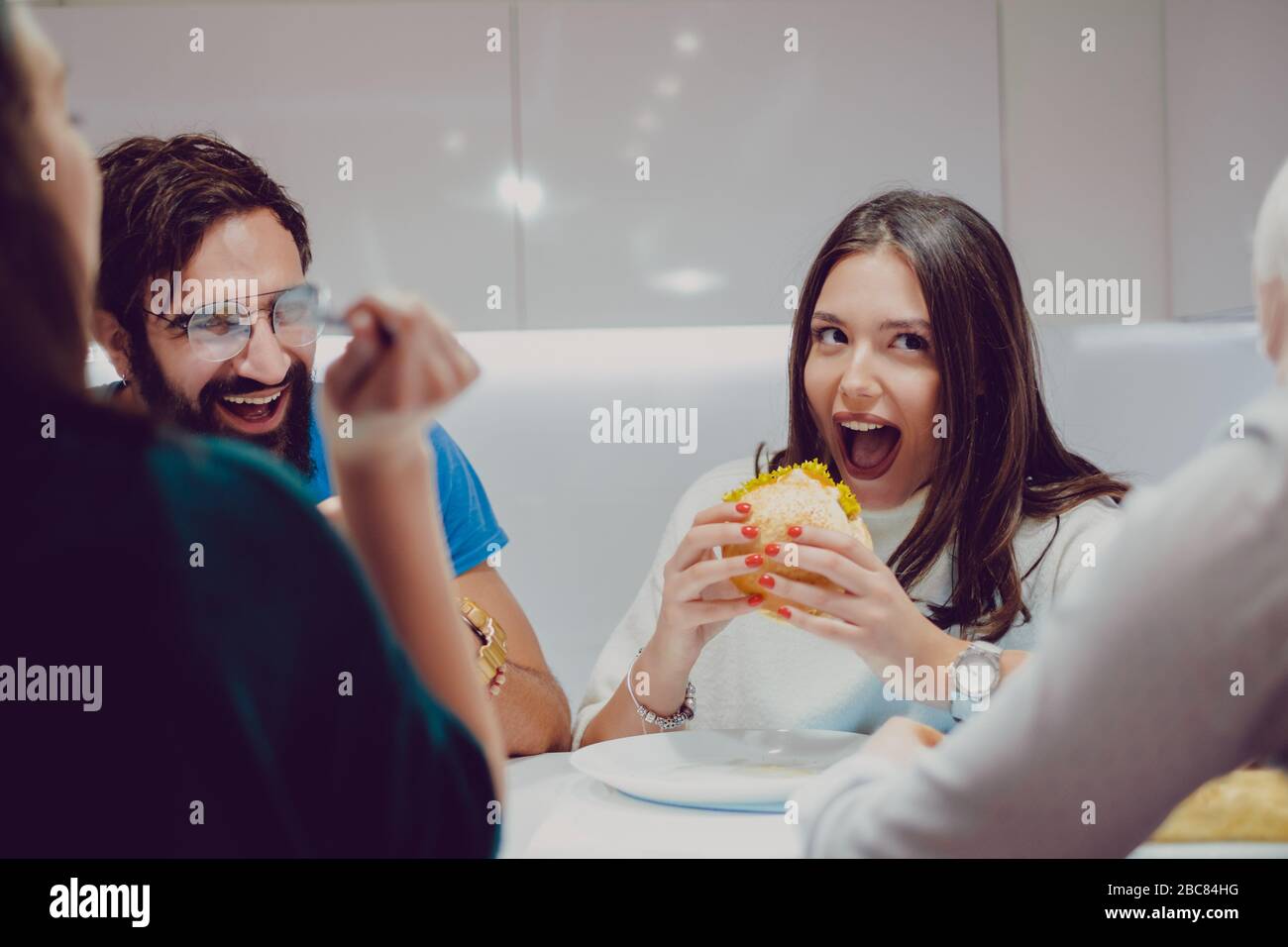 Beautiful brunette girl taking a bite of burger while friends are laughing Stock Photo
