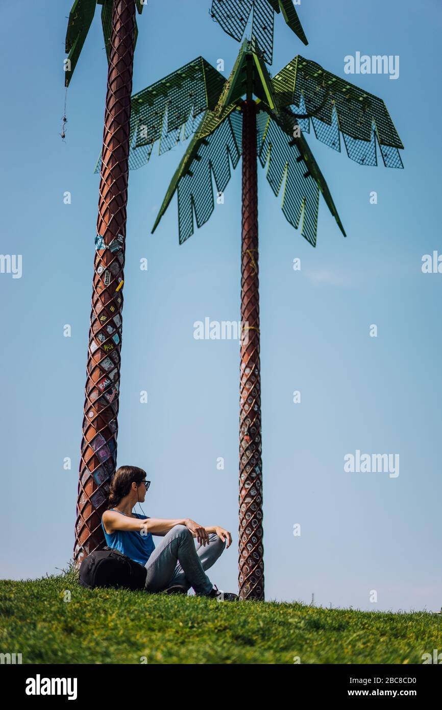 Exploring Hamburg city. Attractive young female tourist with sun glasses sitting under artificial green metal palms in Park. Stock Photo