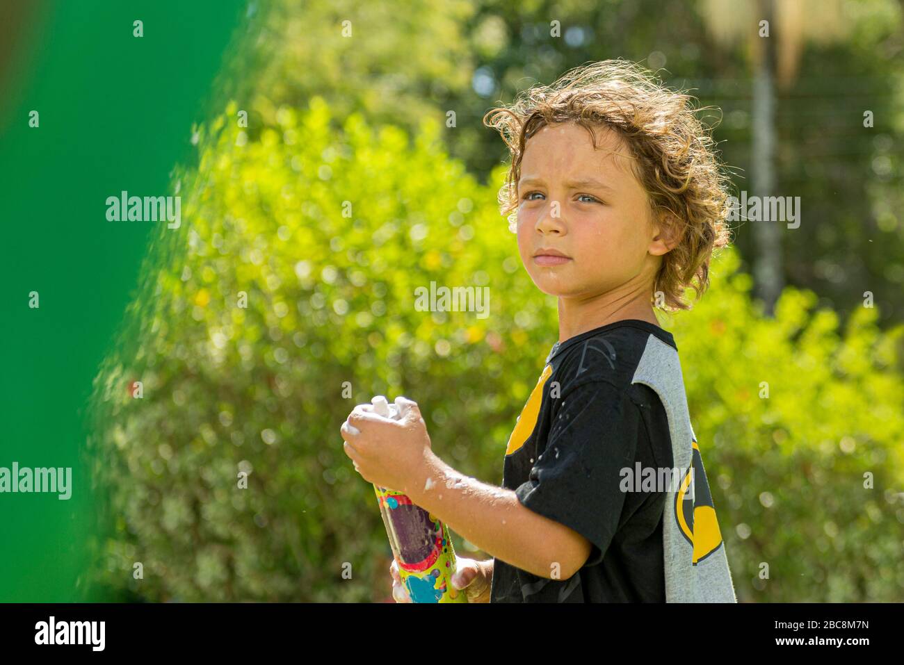 Handsome caucasian boy with curly hair having fun with a can of foam at the carnival outdoors on a beautiful sunny day with blurred vegetation in the Stock Photo