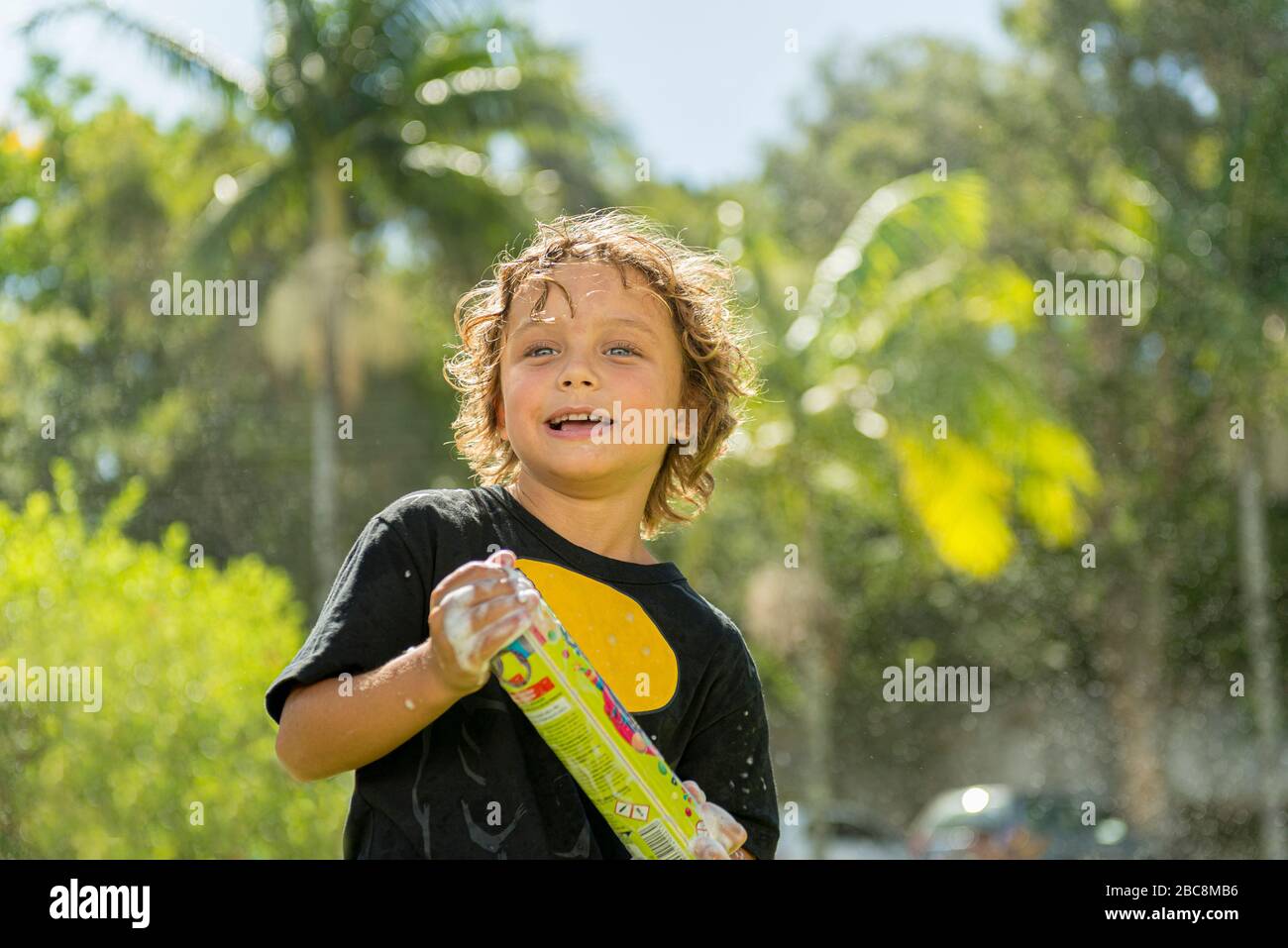 Handsome caucasian boy with curly hair having fun with a can of foam at the carnival outdoors on a beautiful sunny day with blurred vegetation in the Stock Photo